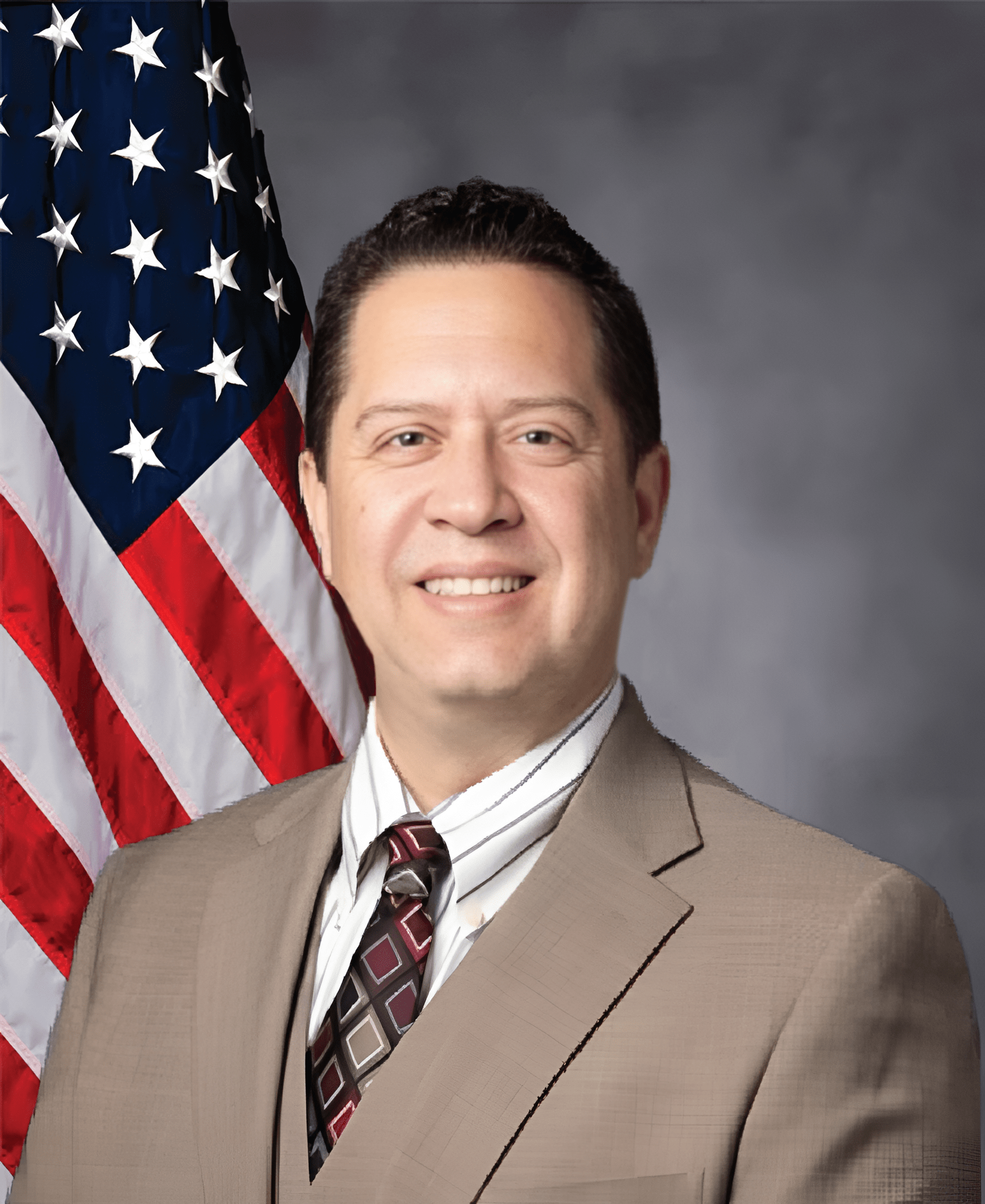 Man in a tan suit and striped tie smiles confidently in front of the American flag, representing a formal or governmental context.