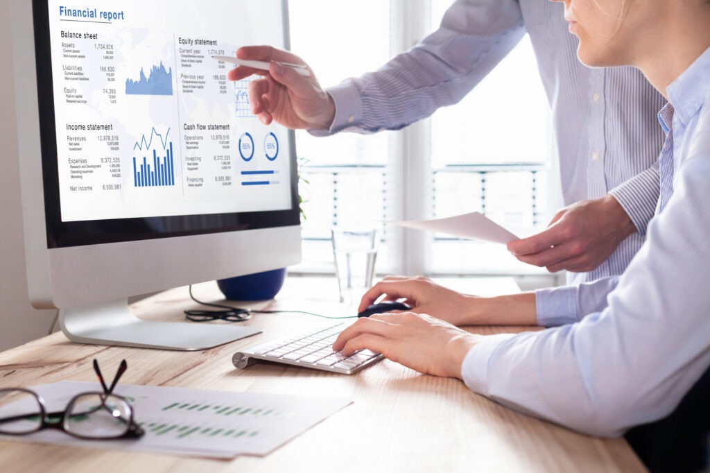 Two professionals review a financial report on a monitor, discussing metrics such as balance sheets and cash flow statements. Documents are on the desk.
